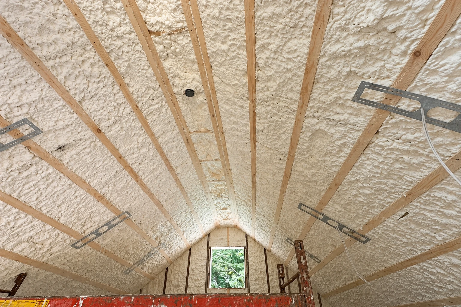 White spray foam covers the sloped ceiling between wooden rafters in an unfinished attic.
