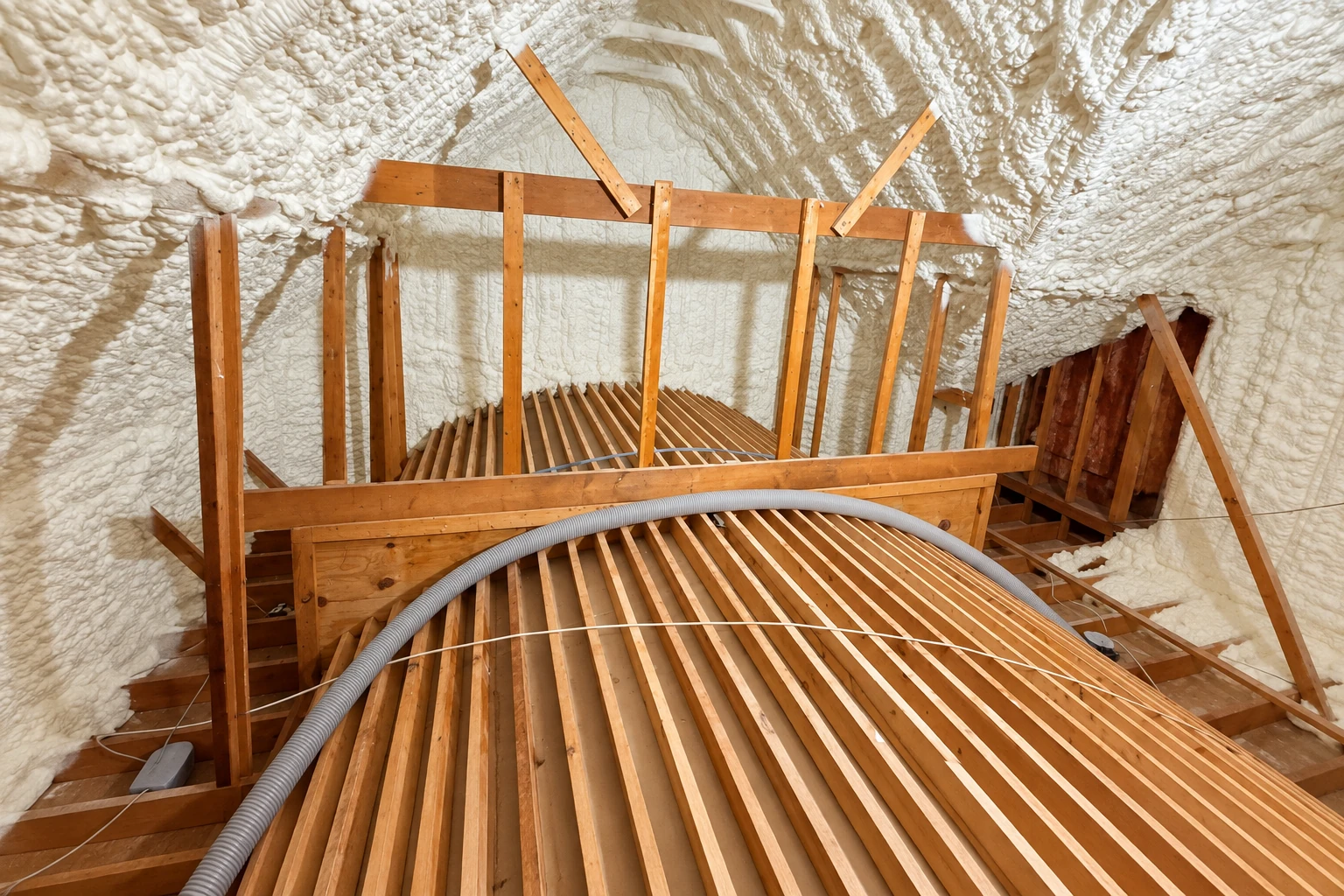 Attic roof coated in white spray foam above exposed wooden framing and a slatted floor.