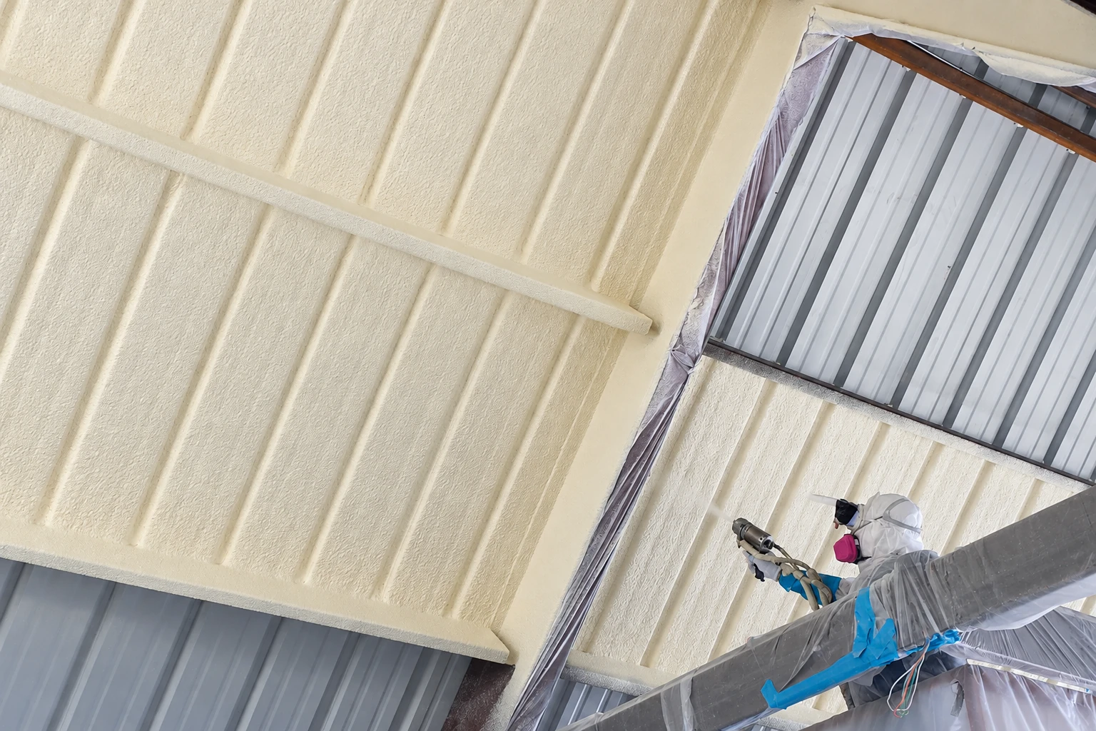 A worker in protective gear sprays cream-colored foam insulation onto the ribbed metal ceiling of a commercial building.