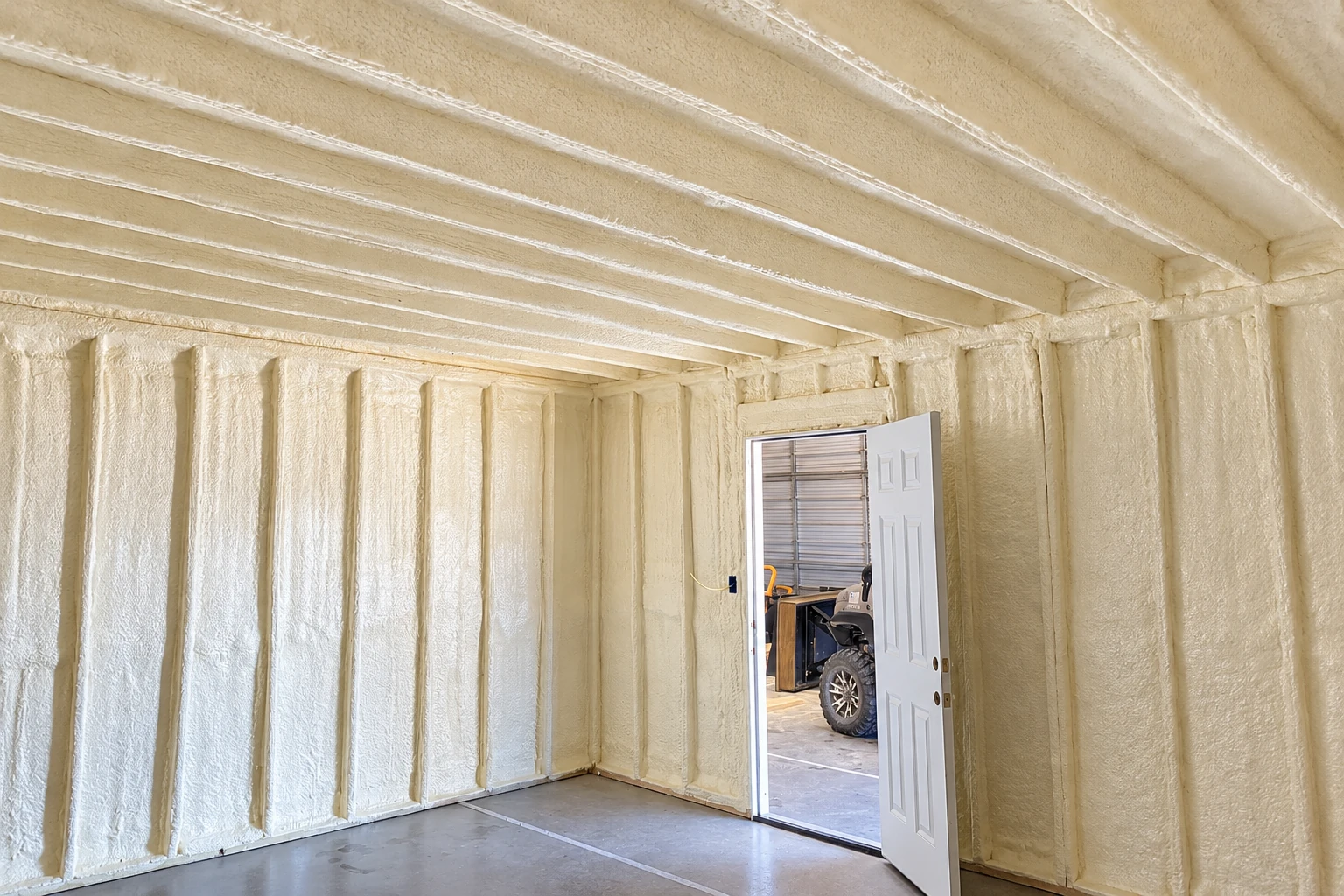 A garage interior with pale spray foam covering the walls and ceiling around the framing, beside an open door.