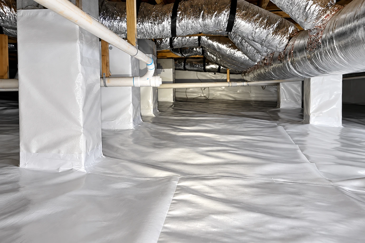 A crawl space floor and support piers are covered in white vapor barrier sheeting beneath metal ducts and pipes.