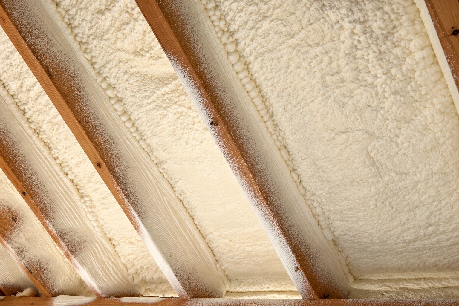 Cream-colored spray foam fills the spaces between exposed wooden rafters.
