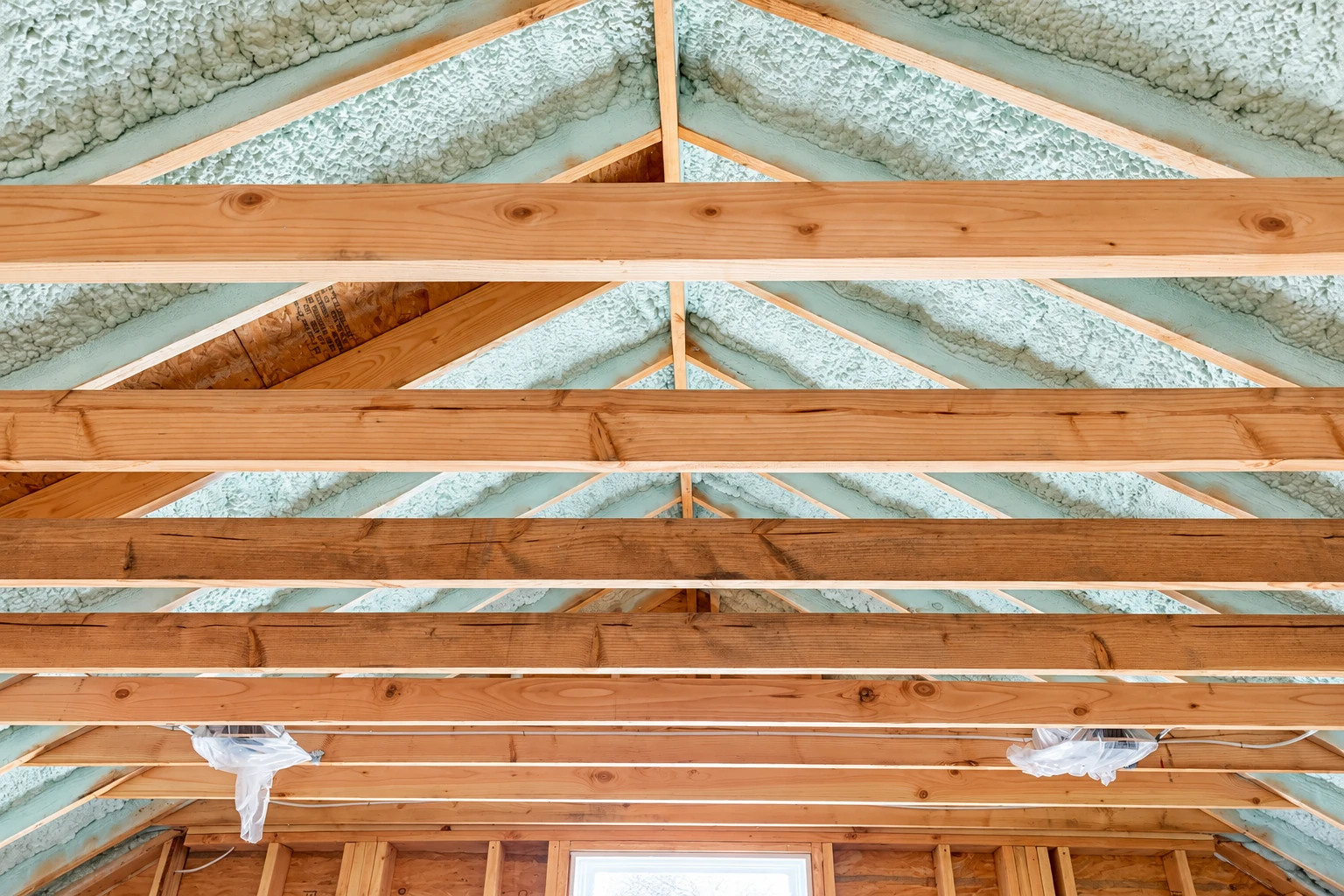 White spray foam insulation fills the roofline between exposed wooden attic trusses.